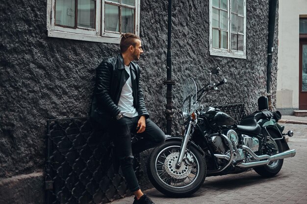 Modischer Biker in schwarzer Lederjacke und Jeans, der sich an einer Wand in der Nähe seines Retro-Motorrads auf einer alten europäischen Straße lehnt.