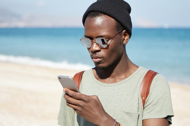 Modischer ernsthafter afrikanischer Mann-Rucksacktourist, der Bilder über soziale Medien unter Verwendung der 3g- oder 4g-Internetverbindung auf dem Mobiltelefon postet, während er um die Welt reist, blauer Ozean und Himmel im Horizont