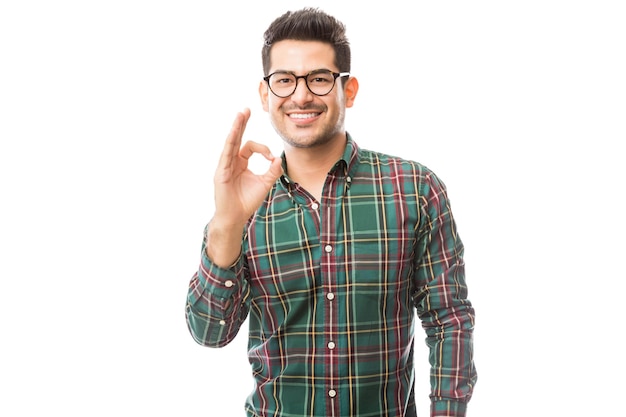 Modischer junger Mann, der OK-Geste zeigt, während er über normalem Hintergrund lächelt