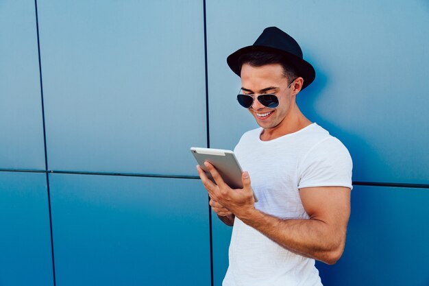 Modischer junger Mann in der Sonnenbrille und im Hut, unter Verwendung einer Tablette, stehend gegen die Wand