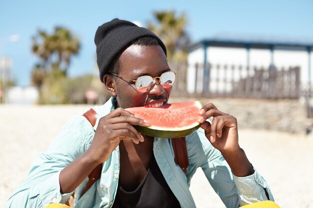 Modischer junger Reisender, der reife saftige Wassermelone beißt, während er tagsüber am Meer entspannt