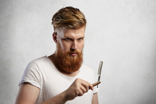 Modischer rothaariger Friseur mit stilvollem Haarschnitt und flockigem Bart, der in seinen Händen ein Rasiermesser mit ernstem Gesichtsausdruck hält.