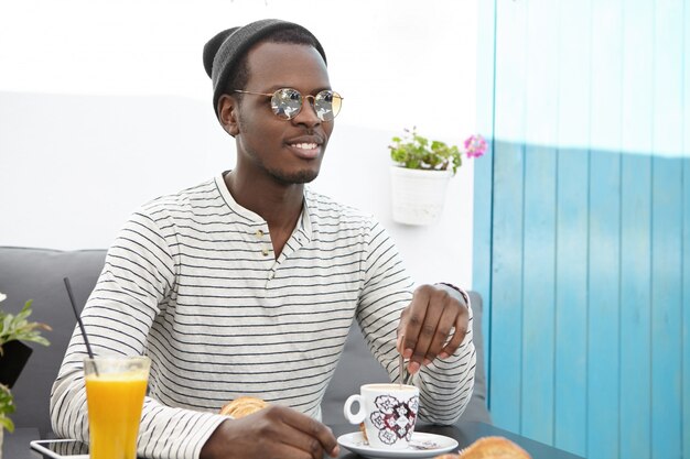 Modischer schwarzer Mann in der runden Sonnenbrille, im gestreiften Hemd und in der Kopfbedeckung, die im Straßencafé ruhen, Kaffee genießen, fröhlich aussehen, sich während der Reise in fremdes Land entspannt und sorglos fühlen