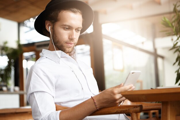 Modischer Student mit weißen Kopfhörern, die kostenloses WLAN nutzen, um seinen Freund auf seinem Handy per Video anzurufen und auf den Bildschirm zu schauen und zu lächeln. Junger Hipster in der schwarzen Kopfbedeckungsnachricht online