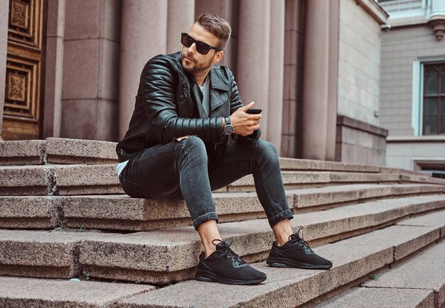 Modischer Typ in schwarzer Jacke und Jeans hält ein Smartphone, das auf Stufen gegen ein altes Gebäude in Europa sitzt.