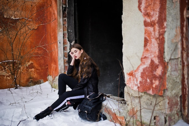 Modisches brünettes Modell mit langen Beinen im langen schwarzen Mantel posierte im Freien am Wintertag gegen die alte Grunge-Wand