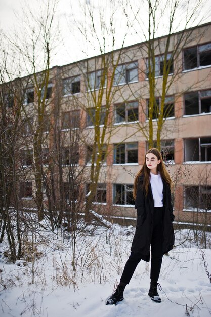 Modisches brünettes Modell mit langen Beinen im langen schwarzen Mantel posierte im Freien am Wintertag gegen verlassene Fabrik