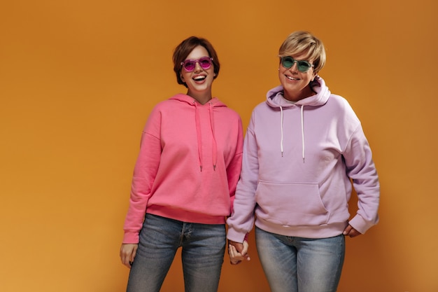 Modisches junges brünettes Mädchen in der rosa Sonnenbrille und im Kapuzenpulli, die alte Dame in den grünen Gläsern auf orange Hintergrund lachen und Hand halten.