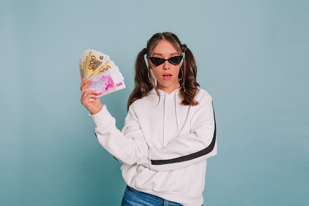 Kostenloses Foto modisches mädchen mit gesammeltem haar, das schwarze sonnenbrille und weißen pullover trägt, der mit einem bündel geld auf isolierter wand aufwirft