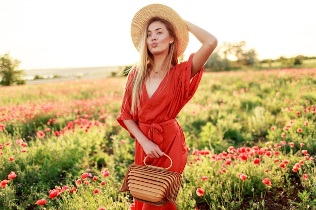 Modisches Porträt im Freien der atemberaubenden blonden Frau, die während des Spaziergangs im erstaunlichen Mohnfeld im warmen Sommerabend aufwirft. Tragen von Strohhut, trendiger Tasche und rotem Kleid.