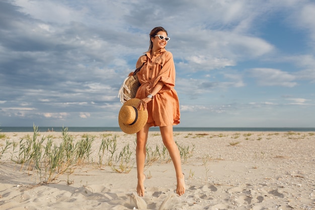 Modisches Sommerbild der schönen Brünettenfrau im trendigen Leinenkleid, das Strohsack hält. Hübsches schlankes Mädchen, das Wochenenden in der Nähe des Ozeans genießt.