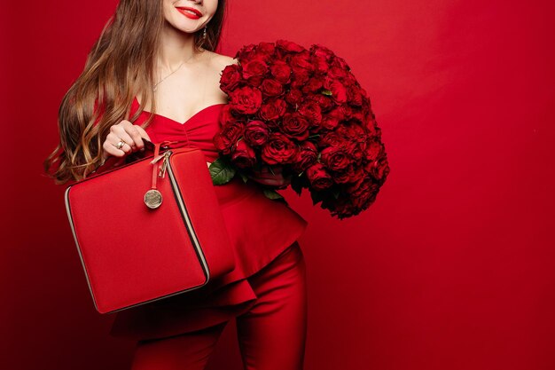 Modisches Studioporträt der stilvollen jungen Frau mit langen braunen Haaren im teuren roten Anzug mit roter Ledertasche und Strauß roter Rosen Sie lächelt in die Kamera Rote Lippen Roter Hintergrund