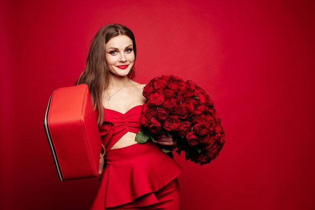 Modisches Studioporträt der stilvollen jungen Frau mit langen braunen Haaren im teuren roten Anzug mit roter Ledertasche und Strauß roter Rosen Sie lächelt in die Kamera Rote Lippen Roter Hintergrund