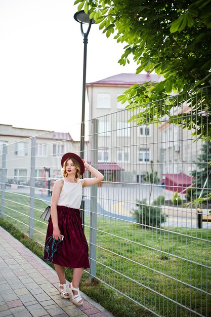 Modisches und schönes blondes Model-Mädchen in stylischem rotem Samt-Velours-Rock, weißer Bluse und Hut posiert mit Telefon und Kopfhörern