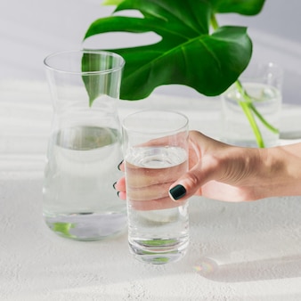 Morgenglas und glas sauberes wasser mit zwei großen grünen blättern auf weißem tisch. frauenhand hält ein glas. frühstück mit langem schatten und reflexion des sonnenlichts. konzept eines gesunden lebensstils
