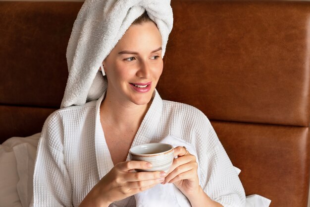 Morgenporträt einer hübschen Frau, die sich nach der Dusche im Bett entspannt, Bademantel und Handtuch auf dem Kopf trägt und leckeren Tee trinkt