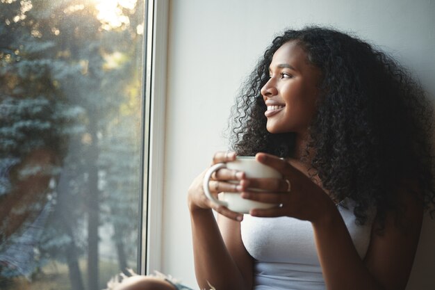 Morgenroutine. Porträt der glücklichen bezaubernden jungen Mischlingsfrau mit gewelltem Haar, die Sommerblick durch Fenster genießt, guten Kaffee trinkt, auf Fensterbank sitzt und lächelt. Schöner Tagträumer