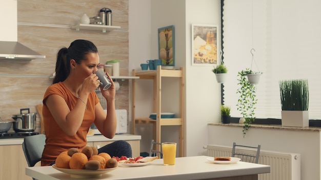 Morgens einen Schluck Kaffee trinken und dabei ein leckeres Frühstück genießen. Dame, die morgens eine Tasse Kaffee genießt. Glückliche Hausfrau entspannt sich und verwöhnt sich allein mit einer gesunden Mahlzeit