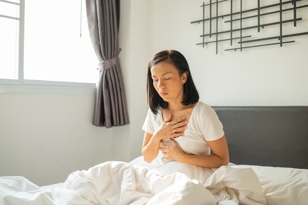 Morgenübelkeit. Junge schwangere Frau, die auf Bett sitzt und ihren Mund bedeckt, der sich während der Schwangerschaft übel fühlt, Frau im weißen Pyjama, der unter saurem Reflux leidet, während sie morgens auf ihrem Bett aufwacht.