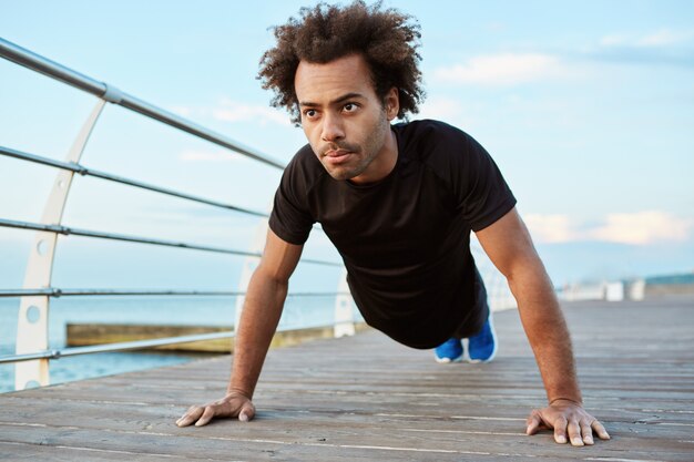Motivierter und konzentrierter afroamerikanischer Athlet mit buschigem Haar, der schwarzes Laufoutfit trägt, das in Plankenposition auf einer hölzernen Plattform steht.