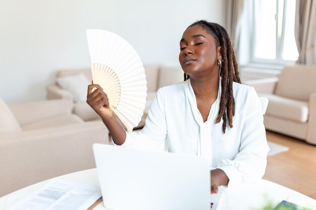 Müde Afrikanerin, die zu Hause am Laptop arbeitet, Welle mit Handventilator, leidet unter Hitzschlag im Innenbereich