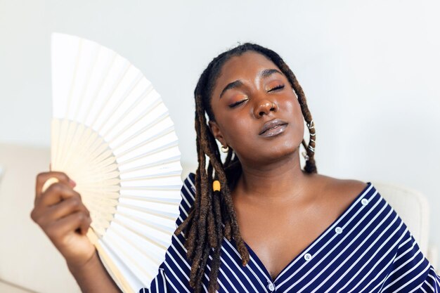 Müde Afro-Frau, die an Hitzschlag oder heißer Sommerwohnung ohne Klimaanlage leidet und einen wehenden Ventilator verwendet, der zu Hause im Wohnzimmer sitzt Überhitzung