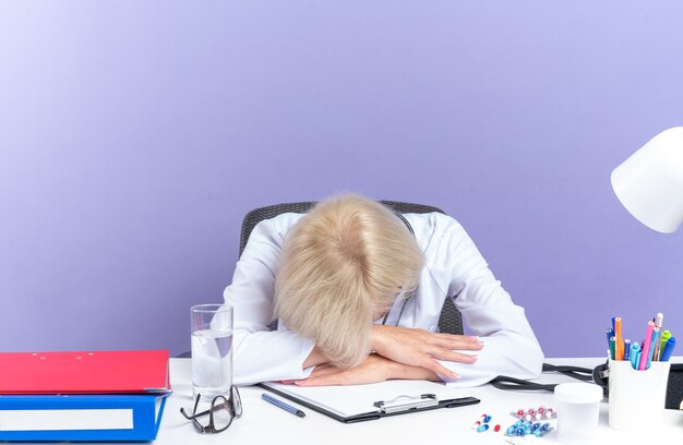 müde erwachsene Ärztin in medizinischer Robe mit Stethoskop am Schreibtisch sitzend mit Bürowerkzeugen, die den Kopf auf den Schreibtisch legen, isoliert auf lila Wand mit Kopienraum