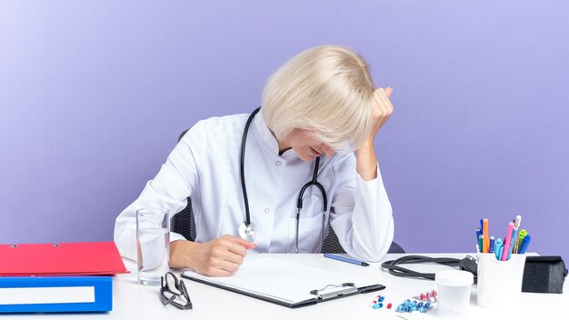 Müde erwachsene slawische Ärztin in medizinischer Robe mit Stethoskop am Schreibtisch sitzend mit Bürowerkzeugen, die Hand auf die Stirn legen, isoliert auf lila Wand mit Kopierraum