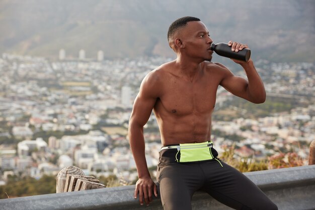 Müde Fitness dunkelhäutige sportliche Person trinkt Wasser, verhindert sich vor Austrocknung, hält Sportflasche, Müdigkeit nach Sport Sommer Übung, führt aktiven gesunden Lebensstil, sitzt am Straßenschild