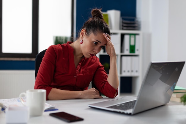 Müde Geschäftsfrau, die am Startgeschäftsschreibtisch sitzt und auf den Laptopbildschirm schaut. Unternehmer im roten Hemd mit Kopfschmerzen im Büro. Kleinunternehmer mit Schwierigkeiten.