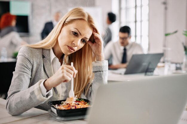 Müde Geschäftsfrau, die sich den Kopf vor Schmerzen hält, während sie etwas auf dem Laptop liest und Gemüsesalat im Büro isst