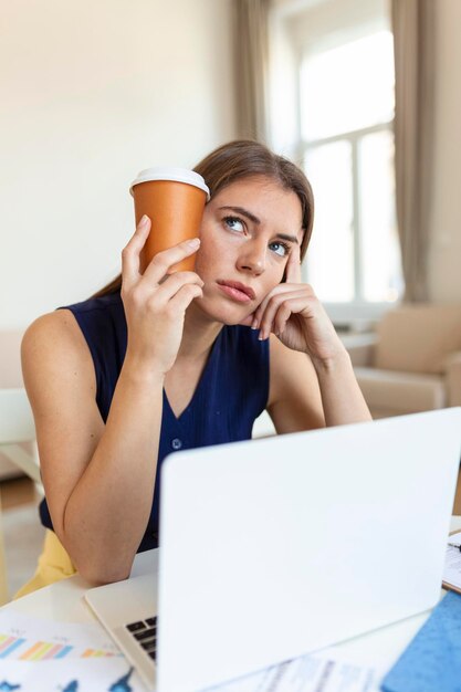 Müde junge Frau, die wegschaut und eine Tasse Kaffee in der Nähe des Kopfes im Büro auf verschwommenem Vordergrund hält