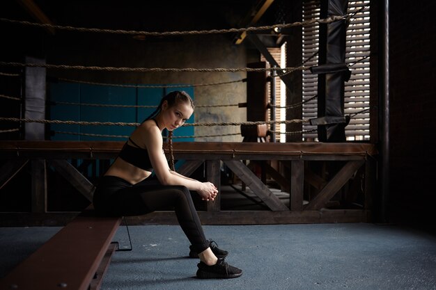 Müde junge Frau mit schlankem Körper, der nach dem Boxtraining im modernen Fitnessstudio auf Bank sitzt und schwarzes Sportoutfit und Turnschuhe trägt