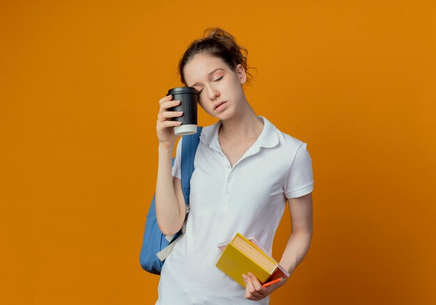 Müde junge hübsche Studentin, die Rückentasche hält, die Buchnotizstift hält und Auge mit Plastikkaffeetasse mit geschlossenen Augen lokalisiert auf orange Hintergrund mit Kopienraum berührt