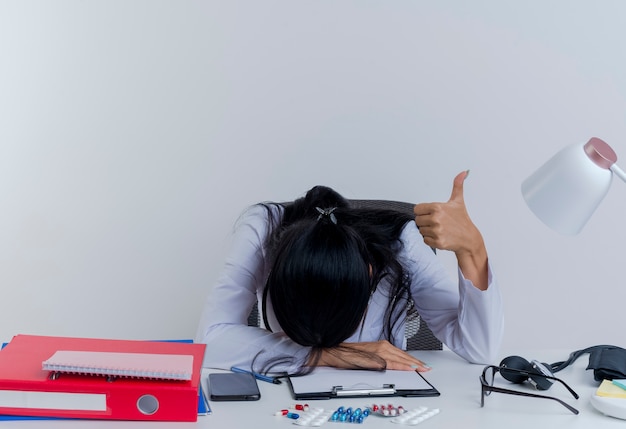 Müde junge Ärztin, die medizinische Robe und Stethoskop trägt, sitzt am Schreibtisch mit medizinischen Werkzeugen, die Hand auf Schreibtisch setzen und Kopf auf Hand zeigen Daumen oben isoliert