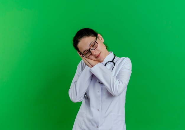 Müde junge Ärztin, die medizinische Robe und Stethoskop und Brille trägt, die Schlafgeste mit geschlossenen Augen tut, lokalisiert auf grüner Wand mit Kopienraum