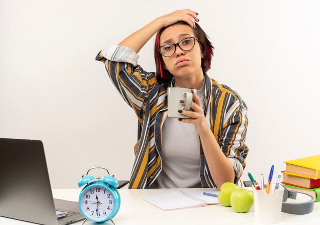 Müde junge Studentin, die eine Brille trägt, die am Schreibtisch mit Universitätswerkzeugen sitzt, die Hand auf Kopf setzen und Tasse Kaffee lokalisiert auf weißem Hintergrund halten