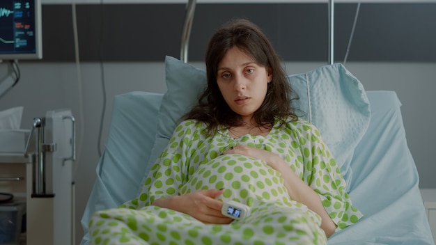 Müde kaukasierin mit schwangerschaftsbauch sitzt im bett im mutterschaftszimmer. junge schwangere person mit oximeter, die spannung über die geburt in der krankenstation verspürt. erwachsener bereitet sich auf die geburt vor Kostenlose Fotos