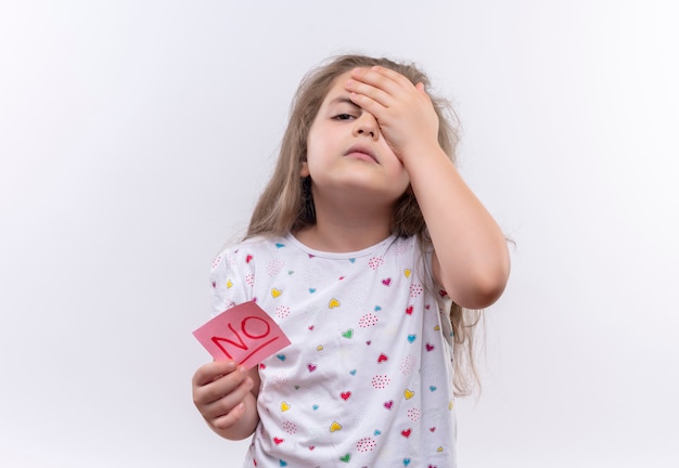 Müde kleine Schulmädchen, die weißes T-Shirt mit Papiermarke tragen, legte ihre Hand auf Stirn auf isolierte weiße Wand