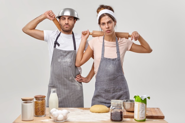 Kostenloses Foto müde köche tragen schürzen, halten nudelhölzer hinter dem rücken, machen nach dem teig eine pause, beschäftigen sich mit der menüplanung und posieren in der küche. kulinarische partner bereiten gemeinsam hausgemachten kuchen für besondere anlässe zu