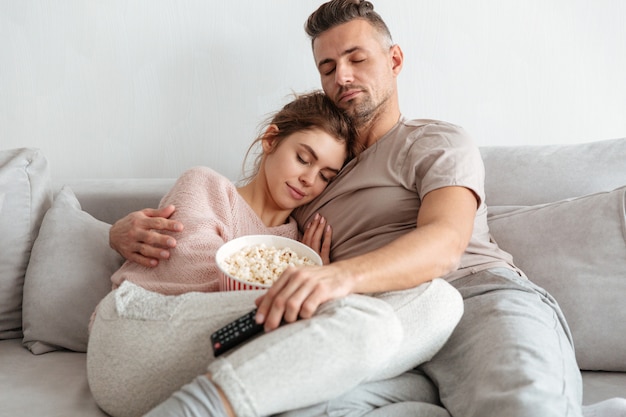 Müde liebende Paare, die auf Couch zusammen mit Popcorn sitzen und zu Hause Rest haben