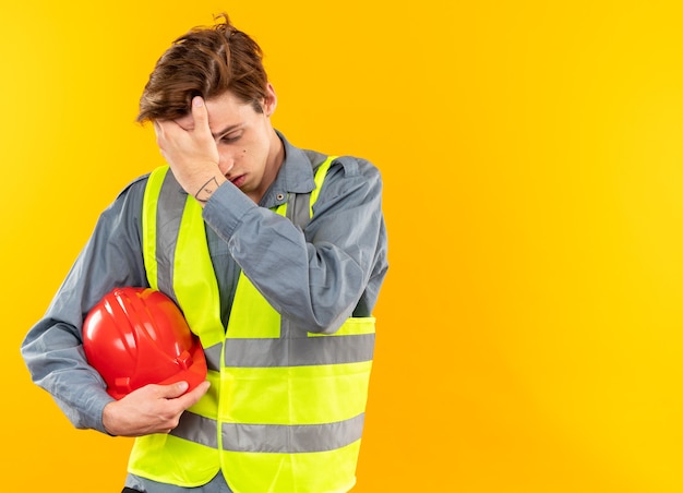 Müde mit gesenktem Kopf junger Baumeister in Uniform mit Schutzhelm, der die Hand auf die Stirn legt, isoliert auf gelber Wand