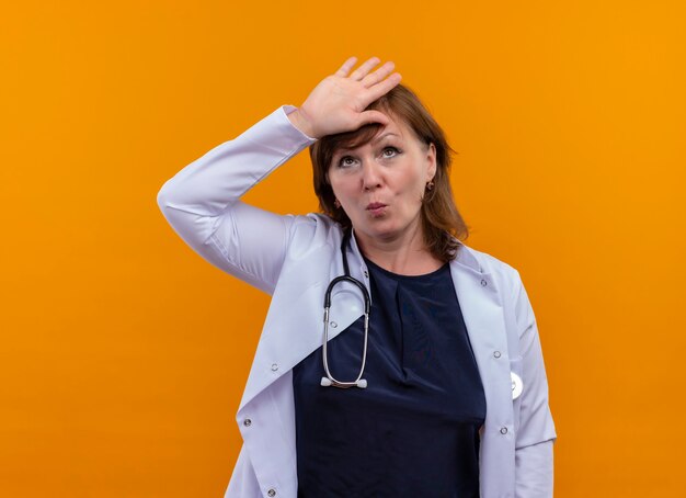Müde Ärztin mittleren Alters, die medizinische Robe und Stethoskop trägt Hand auf Stirn auf isolierte orange Wand mit Kopienraum setzt