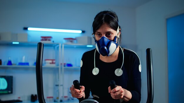 Müde Sportlerin, die auf Crosstrainer trainiert, erhöht den Rhythmus der Übungen mit Maske und medizinischen Elektroden, die die Muskelausdauer und die Herzfrequenz im wissenschaftlichen Sportlabor überwachen monitoring