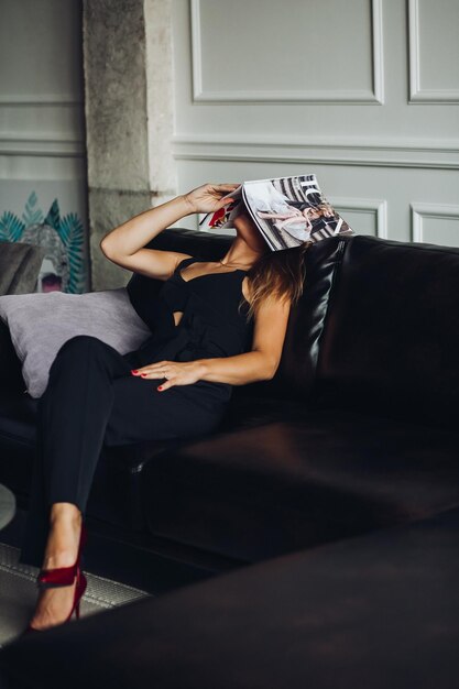 Müde und erschöpfte Frau, die auf einer schwarzen Couch sitzt und das Magazin auf den Kopf legt Schlankes Modell in schwarzem, elegantem Gesamtkonzept der Vogue-Modeindustrie