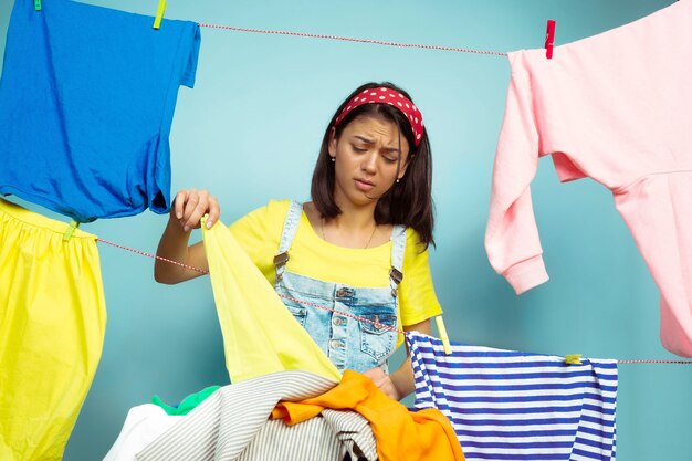 Müde und schöne Hausfrau, die Hausarbeit lokalisiert auf blauem Hintergrund tut. Junge kaukasische Frau, die durch gewaschene Kleidung umgeben ist. Häusliches Leben, helle Kunstwerke, Haushaltskonzept. Sieht verärgert aus.