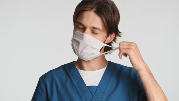 Müder Arzt, der die medizinische Maske nach einem anstrengenden Tag im Krankenhaus schläfrig macht Junger Praktikant in Uniform im Studio Überarbeiteter Ausdruck