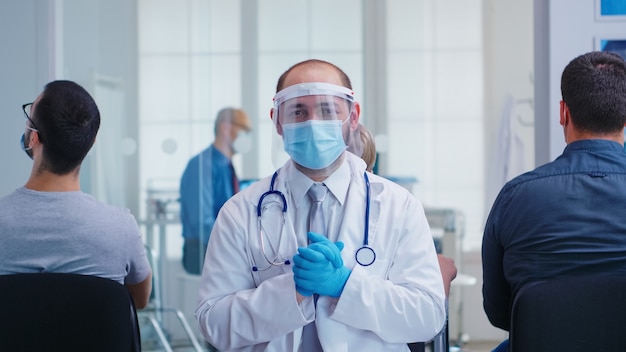 Müder Arzt mit Gesichtsmaske und Visier gegen Coronavirus im Wartebereich des Krankenhauses mit Blick auf die Kamera. Älterer Mann und Krankenschwester im Krankenhausuntersuchungsraum.