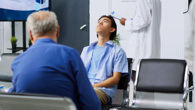 Kostenloses Foto müder asiatischer patient ist während des kontrolltermins nervös, während er darauf wartet, dass der spezialist die medikamentöse behandlung bespricht. junger erwachsener sitzt auf einem stuhl im wartebereich des krankenhauses.