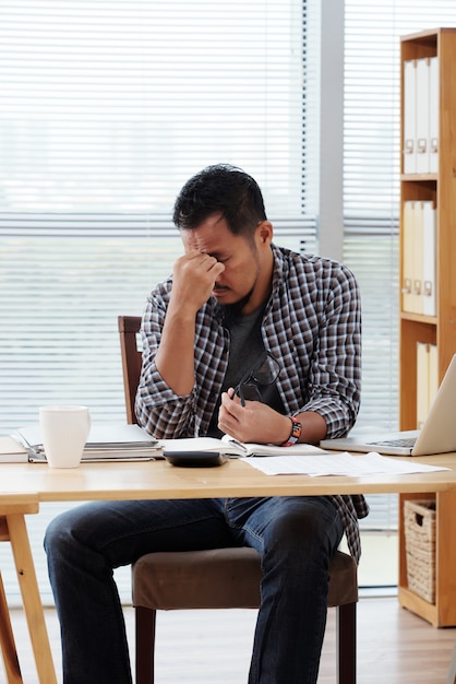 Müder asiatischer Unternehmer, der bei Tisch im Büro sitzt und seine Stirn reibt